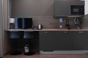 A modern kitchen workspace featuring dark cabinets with a wood countertop. On the left sits a small desk setup with a flat-screen monitor, a lamp, two black chairs, a telephone, and an electric kettle. Above the counter is a wall-mounted microwave and a range hood. Various kitchen utensils hang on a rail beside the sink on the right.