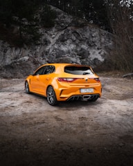 A vibrant orange hatchback car is parked on a rough, natural terrain with rocky cliffs and sparse trees in the background. The car is facing towards the left side of the image, displaying its sleek rear design and sporty features. The setting appears to be an outdoor area, possibly a rural or mountainous region.