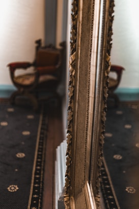 An ornate, gilded mirror reflects part of an elegant room, highlighting the intricate carvings on its frame. In the background, an antique chair with a wooden frame and a richly upholstered backrest is visible. The floor is covered with a dark patterned carpet, adding to the vintage atmosphere of the space.