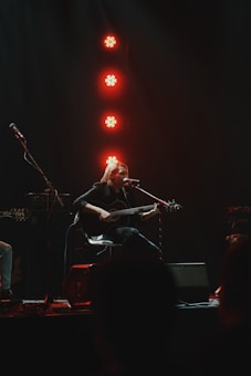 A musician plays an acoustic guitar while seated on stage, illuminated by red stage lights in a dimly lit setting.