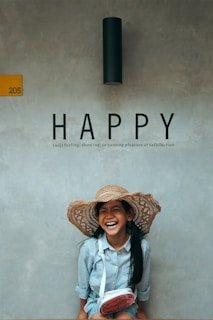 A child with long hair, wearing a large straw hat and a striped shirt, is sitting against a concrete wall. They are laughing joyfully, and there is a yellow sign with the number 205 beside them. Above, the word 'HAPPY' is prominently displayed along with its definition.