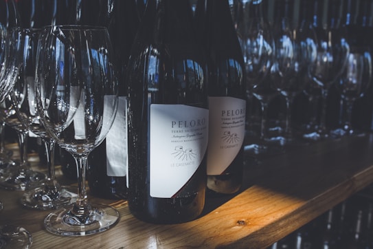 Several wine bottles labeled 'Peloro' are placed on a wooden shelf, surrounded by a row of clear wine glasses. The scene is illuminated by soft, natural light creating a cozy and inviting atmosphere.