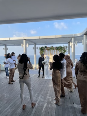 A wedding ceremony taking place on a wooden terrace overlooking the sea, with a couple standing under a floral arch. The bride is in a white dress, and the groom is in a dark suit. Several photographers and guests are gathered around, some dressed in formal wear, capturing the moment.