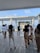 A wedding ceremony taking place on a wooden terrace overlooking the sea, with a couple standing under a floral arch. The bride is in a white dress, and the groom is in a dark suit. Several photographers and guests are gathered around, some dressed in formal wear, capturing the moment.