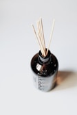 A dark glass bottle with sticks emerging from its opening, creating a minimalist aesthetic against a plain white background. The bottle has a measurement label on its side, and the contents are not visible, highlighting the simplicity and elegance of the design.