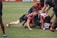 A group of rugby players engaged in a scrum on a grassy field, wearing colorful striped uniforms with matching socks. Some players are on the ground while others are bent over, actively participating in the game. The players show a mix of intense focus and physical exertion.