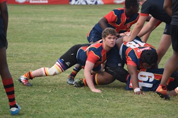 A group of rugby players engaged in a scrum on a grassy field, wearing colorful striped uniforms with matching socks. Some players are on the ground while others are bent over, actively participating in the game. The players show a mix of intense focus and physical exertion.