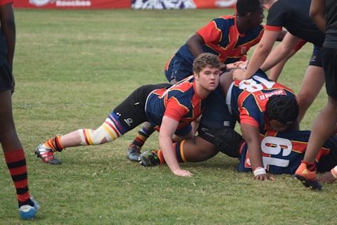 A group of rugby players engaged in a scrum on a grassy field, wearing colorful striped uniforms with matching socks. Some players are on the ground while others are bent over, actively participating in the game. The players show a mix of intense focus and physical exertion.