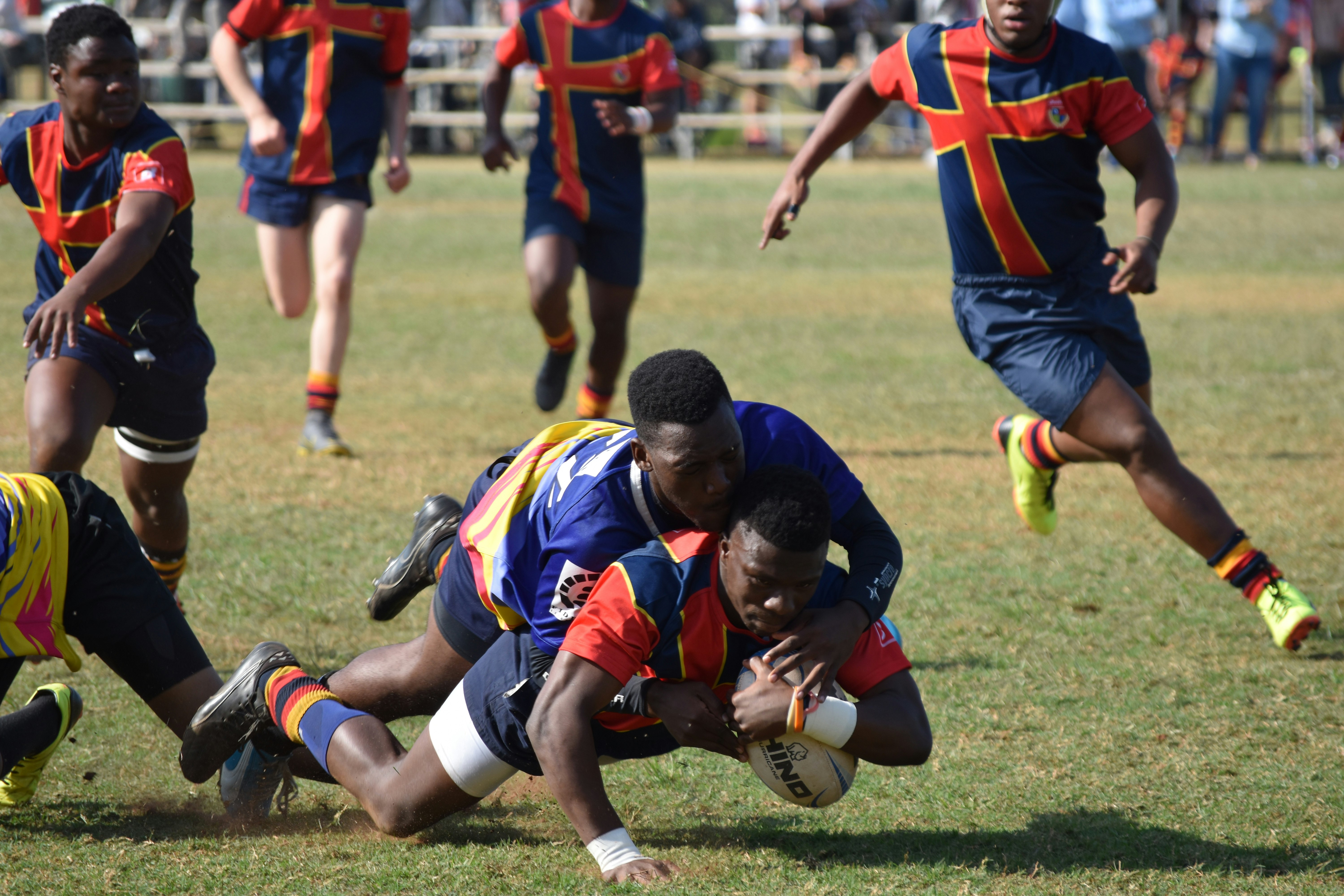 A young man (blue and yellow shirts) tackles his opponent (blue and red shirts) to the ground during a club rugby match in South Africa while the youngster clutches the ball and refuses to let go. 