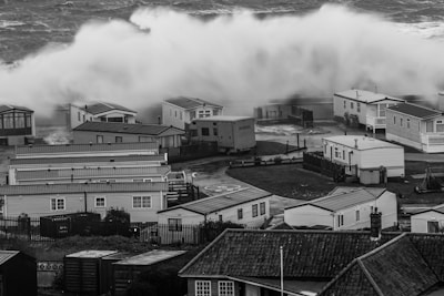 A large wave crashes onto a set of mobile homes near the shoreline, creating a dramatic and intense scene. The area appears to be a residential park with closely packed houses. The wave is powerful, creating a mist as it thunders down. The image is in black and white, enhancing the stark contrast between the wave and the homes.