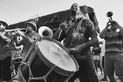 A group of musicians perform energetically, playing drums and brass instruments, with a festive and lively atmosphere. People can be seen in the background enjoying the performance, and the setting appears to be an outdoor event or festival.