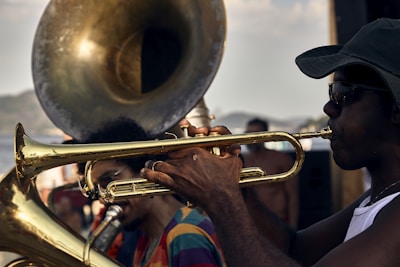Musicians are performing outdoors with brass instruments. A person is playing the trumpet, while another musician, partially visible in the background, is playing the tuba. The setting appears to be lively, with an open-air atmosphere and people gathered around.