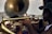 Musicians are performing outdoors with brass instruments. A person is playing the trumpet, while another musician, partially visible in the background, is playing the tuba. The setting appears to be lively, with an open-air atmosphere and people gathered around.