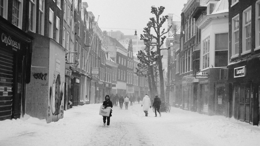 Graustufenfoto von Menschen, die auf einer schneebedeckten Straße zwischen Gebäuden spazieren gehen