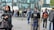 A busy urban street scene with diverse pedestrians. Some individuals wear winter clothing, and there is a visible presence of a police officer with armored gear. A group of people is gathered near a glass-fronted building in the background.