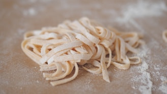 A pile of uncooked flat noodles is resting on a lightly floured surface. The noodles have a pale, slightly yellowish color and are neatly arranged in overlapping loops and twists.