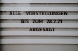 A sign with horizontal slats displays the text 'ALLE VORSTELLUNGEN BIS ZUM 28.2.21 ABGESAGT', indicating that all events are canceled until a specific date. The lettering is bold and clear, set against a muted background.