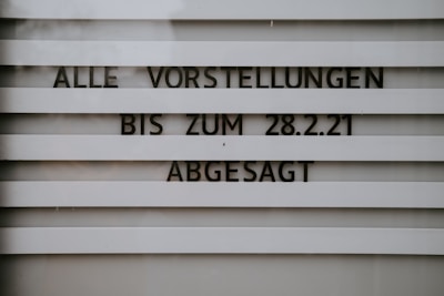 A sign with horizontal slats displays the text 'ALLE VORSTELLUNGEN BIS ZUM 28.2.21 ABGESAGT', indicating that all events are canceled until a specific date. The lettering is bold and clear, set against a muted background.