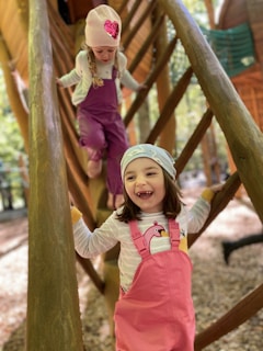 Two young children are climbing on a wooden playground structure. Both are wearing colorful outfits, with one wearing pink overalls featuring a swan design, and the other in purple overalls. The child in the foreground is smiling and wearing a light-colored cap, while the child behind wears a knitted hat with a sequined heart design. The setting appears to be a forested area with autumn leaves on the ground.