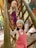 Two young children are climbing on a wooden playground structure. Both are wearing colorful outfits, with one wearing pink overalls featuring a swan design, and the other in purple overalls. The child in the foreground is smiling and wearing a light-colored cap, while the child behind wears a knitted hat with a sequined heart design. The setting appears to be a forested area with autumn leaves on the ground.
