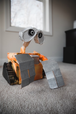 A homemade robot model resembling a character from a popular animated movie is placed on a carpeted floor inside a room. The robot is constructed from cardboard and other recycled materials, with large expressive eyes and grey hands. There is a window in the background letting in natural light.