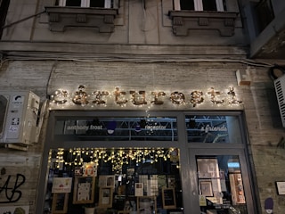 A storefront with illuminated signage displaying the name in decorative lights. The window displays various posters and books, with string lights enhancing the cozy ambiance. The background architecture includes classic elements with a stone facade and decorative moldings. An air conditioning unit is mounted on the left side. The scene is set at night, contributing to a warm and inviting atmosphere.