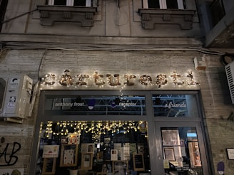 A storefront with illuminated signage displaying the name in decorative lights. The window displays various posters and books, with string lights enhancing the cozy ambiance. The background architecture includes classic elements with a stone facade and decorative moldings. An air conditioning unit is mounted on the left side. The scene is set at night, contributing to a warm and inviting atmosphere.