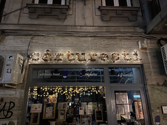 A storefront with illuminated signage displaying the name in decorative lights. The window displays various posters and books, with string lights enhancing the cozy ambiance. The background architecture includes classic elements with a stone facade and decorative moldings. An air conditioning unit is mounted on the left side. The scene is set at night, contributing to a warm and inviting atmosphere.