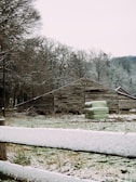 A rustic wooden barn stands amidst a winter landscape. Surrounding trees are bare, dusted with a light layer of snow. In front of the barn, a few large green hay bales are stacked. A wooden fence in the foreground is also covered with snow, adding to the serene, chilly atmosphere.