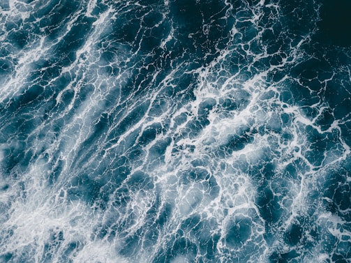 An aerial view of ocean waves creating intricate patterns with white foam on the deep blue water surface.
