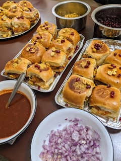 Several trays of stuffed bread rolls topped with pomegranate seeds and nuts are arranged on a table. There are also bowls containing chopped onions and a brown liquid sauce. The scene appears to be a setup for serving food, with an emphasis on Indian-style cuisine.