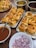 Several trays of stuffed bread rolls topped with pomegranate seeds and nuts are arranged on a table. There are also bowls containing chopped onions and a brown liquid sauce. The scene appears to be a setup for serving food, with an emphasis on Indian-style cuisine.