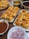 Several trays of stuffed bread rolls topped with pomegranate seeds and nuts are arranged on a table. There are also bowls containing chopped onions and a brown liquid sauce. The scene appears to be a setup for serving food, with an emphasis on Indian-style cuisine.