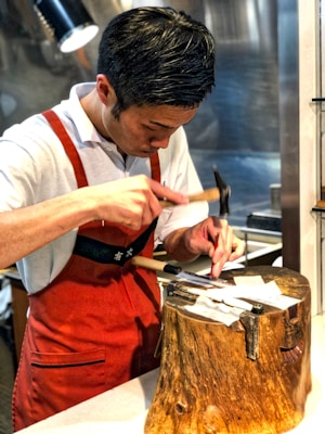 A person is focused on a crafting task, using a small hammer on a metal object placed on a wooden block. They are wearing a red apron over a white shirt, and the setting appears to be a workshop or kitchen environment.