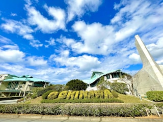 A modern architectural structure with a unique design, featuring a curved roof and extensive use of glass. In the foreground, there is a bush trimmed to form the word 'COMINHA'. The sky is clear with scattered fluffy clouds.