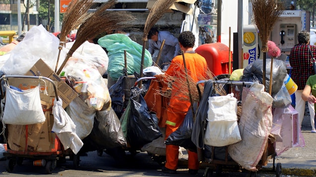 A sanitation worker wearing an orange uniform loads garbage onto a truck. Various carts and bags filled with waste materials like cardboard, plastic, and other refuse surround him. Several brooms are visible, indicating cleaning activities. In the background, people pass by and a telephone booth is visible.