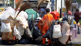 A sanitation worker wearing an orange uniform loads garbage onto a truck. Various carts and bags filled with waste materials like cardboard, plastic, and other refuse surround him. Several brooms are visible, indicating cleaning activities. In the background, people pass by and a telephone booth is visible.