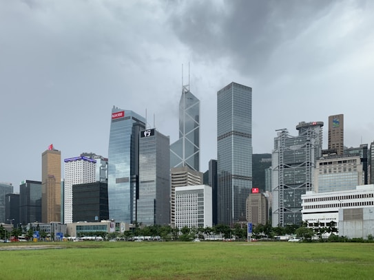 A skyline dominated by modern skyscrapers with various architectural styles, set against a cloudy sky. The foreground features a lush green lawn, creating a contrast with the urban environment. Distinctive logos and markings identify some buildings.