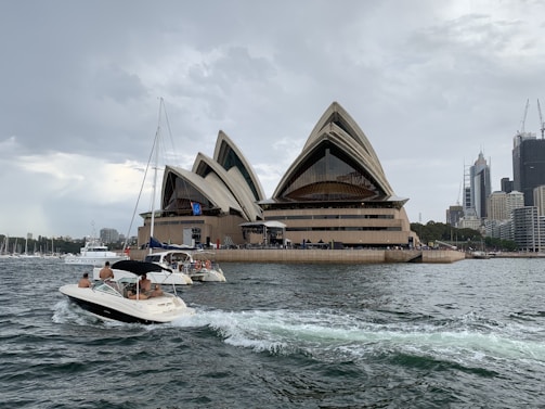 A waterfront scene featuring a famous opera house with its distinctive sail-like architecture. Several boats with people on board are visible on the water, and there's a city skyline with tall buildings in the background under a cloudy sky.