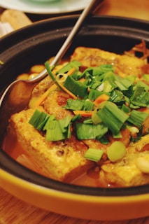 A dish consisting of golden brown tofu blocks topped with vibrant green scallions, served in a dark pot. The tofu sits in a savory sauce, and a metal spoon is resting in the pot, adding a touch of silver contrast.