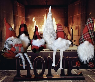 Several whimsical gnome figurines are arranged sitting atop a wooden block sign spelling out 'HOME' in front of a lit fireplace. The gnomes have long, pointed hats in red plaid, buffalo check, and white knit patterns, along with fluffy white beards. One gnome is holding a small plaid-wrapped gift. The background features a warm, glowing fire casting a cozy ambiance.