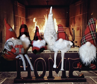 Several whimsical gnome figurines are arranged sitting atop a wooden block sign spelling out 'HOME' in front of a lit fireplace. The gnomes have long, pointed hats in red plaid, buffalo check, and white knit patterns, along with fluffy white beards. One gnome is holding a small plaid-wrapped gift. The background features a warm, glowing fire casting a cozy ambiance.