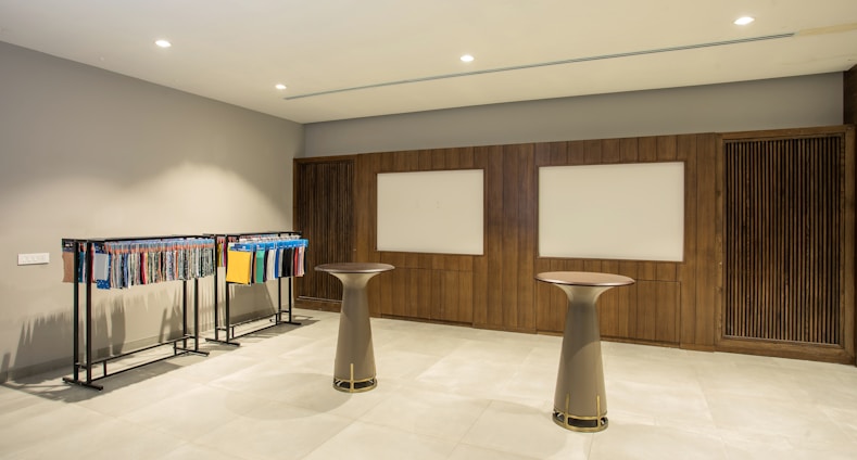 A modern interior space featuring two tall round tables and a clothing rack displaying various fabrics or textile samples in an organized manner. The walls are adorned with wooden paneling and two rectangular whiteboards or display panels. The lighting is bright and evenly distributed, and the floor is covered with tiles.