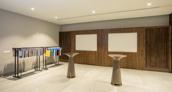 A modern interior space featuring two tall round tables and a clothing rack displaying various fabrics or textile samples in an organized manner. The walls are adorned with wooden paneling and two rectangular whiteboards or display panels. The lighting is bright and evenly distributed, and the floor is covered with tiles.