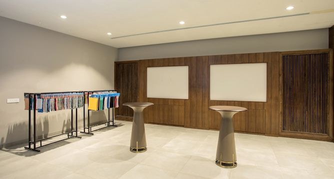 A modern interior space featuring two tall round tables and a clothing rack displaying various fabrics or textile samples in an organized manner. The walls are adorned with wooden paneling and two rectangular whiteboards or display panels. The lighting is bright and evenly distributed, and the floor is covered with tiles.
