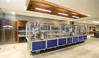 A modern industrial setting featuring an automated assembly line machine enclosed in glass. The room is well-lit with a sleek design, including wooden accents and clean surfaces. There is a focus on the machinery, with components visible inside the enclosure.