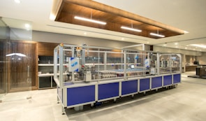 A modern industrial setting featuring an automated assembly line machine enclosed in glass. The room is well-lit with a sleek design, including wooden accents and clean surfaces. There is a focus on the machinery, with components visible inside the enclosure.