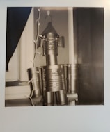 A black and white image of a robot figure assembled from tin cans. It is positioned near a window, and a string of decorative lights is draped beside it. The robot has cylindrical limbs and a cone-shaped head with a hook on top.
