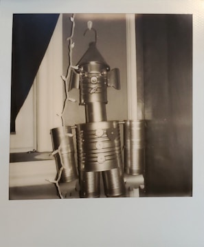 A black and white image of a robot figure assembled from tin cans. It is positioned near a window, and a string of decorative lights is draped beside it. The robot has cylindrical limbs and a cone-shaped head with a hook on top.