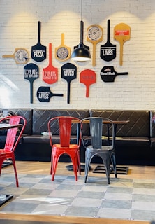 A cozy seating area in a restaurant, featuring a wall decorated with colorful pizza paddles with various inspirational phrases. The seating consists of red and black metal chairs and a black cushioned bench against the wall. A single pendant light hangs above, casting a warm glow on the wall.